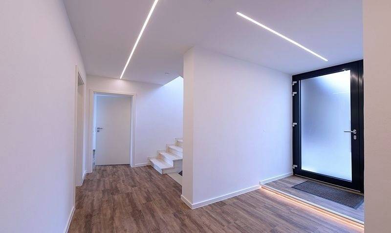 Moderner, minimalistisch gestalteter Flur mit Holzfußboden, LED-Leuchtstreifen an der Decke und Treppe, die zu einer oberen Etage führt.