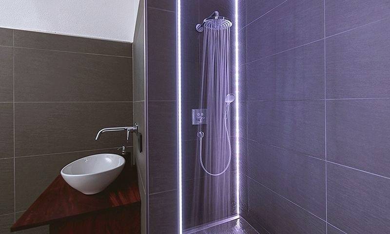 Moderne Dusche mit digitaler Armatur und LED-Beleuchtung in einem Badezimmer mit dunklen Fliesen. Links steht ein weißes Waschbecken.