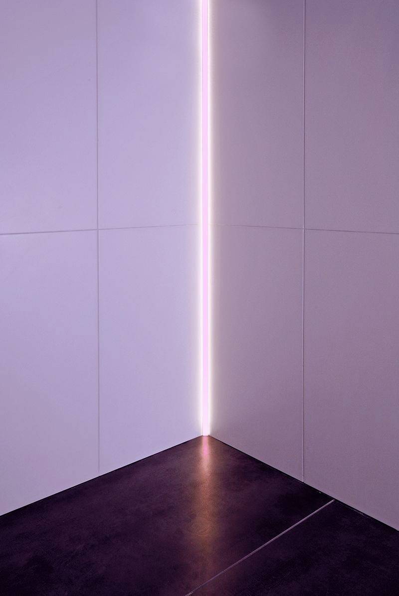 Eine minimalistische Ecke eines Raumes mit weißen Wänden und einem schmalen vertikalen Lichtstreifen in der Mitte. Dunkler Boden.