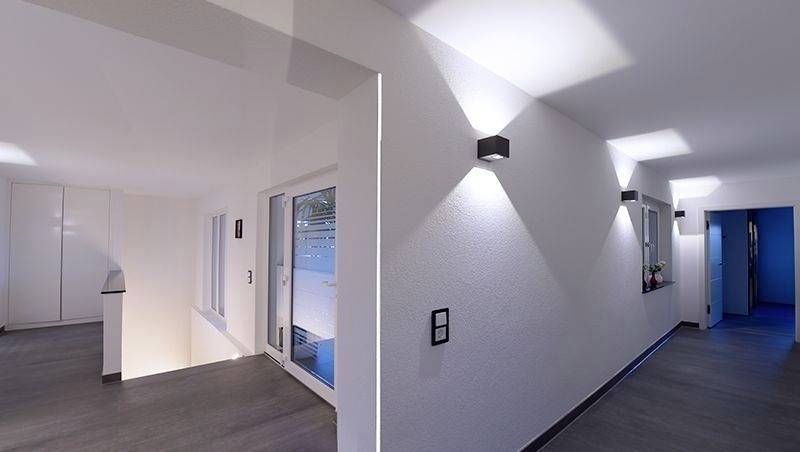 Moderner Innenraum eines Gebäudes mit weiß gestrichenen Wänden und dunklem Holzboden, beleuchtet von stilvollen Wandlampen.