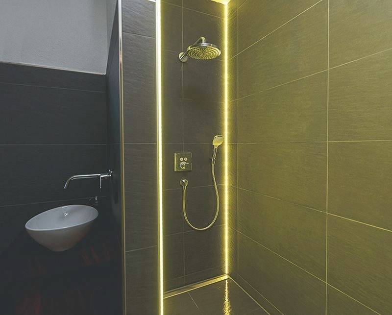 Duschbereich mit moderner Regendusche, beleuchtet durch dezente LED-Lichtleiste; graue Fliesen umgeben die Dusche. Links ein Waschbecken.