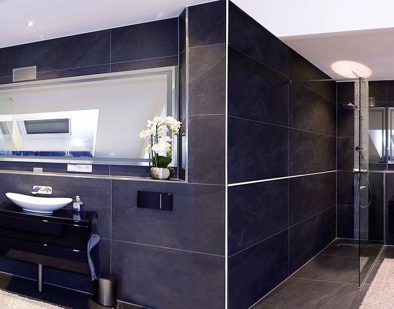 Modernes Badezimmer mit dunklen Fliesen. Links ein Waschbecken mit Spiegel und Orchideen. Rechts eine gläserne Dusche.