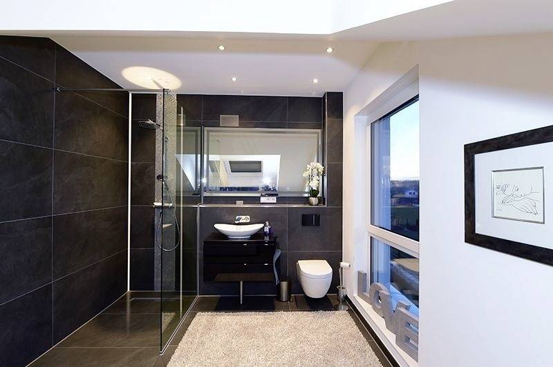 Modernes Badezimmer mit schwarzer Fliesenwand, bodengleicher Dusche und Waschbecken, großer Spiegel, weißer Toilette und Fenster.