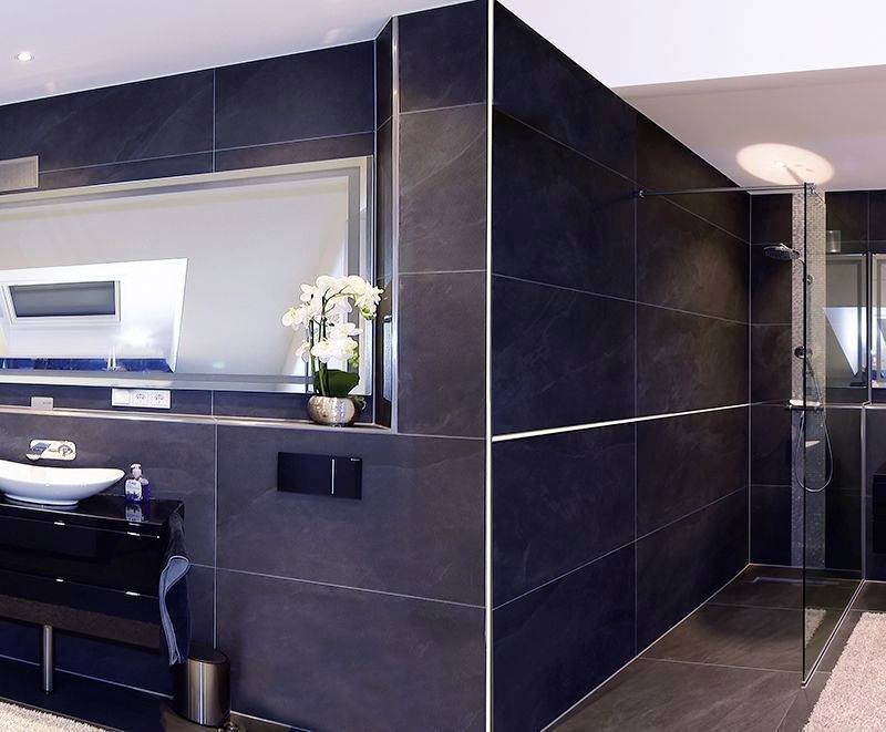 Modernes Badezimmer mit dunklen Fliesen, Waschbecken, großem Spiegel und Orchidee. Dusche mit Glaswand im Hintergrund. Minimalistisches Design.