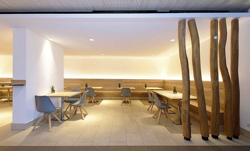 Ein modernes Café mit Holztischen, grauen Stühlen und heller Beleuchtung. Holzsäulen dienen als Raumtrenner, Pflanzen dekorieren die Tische.