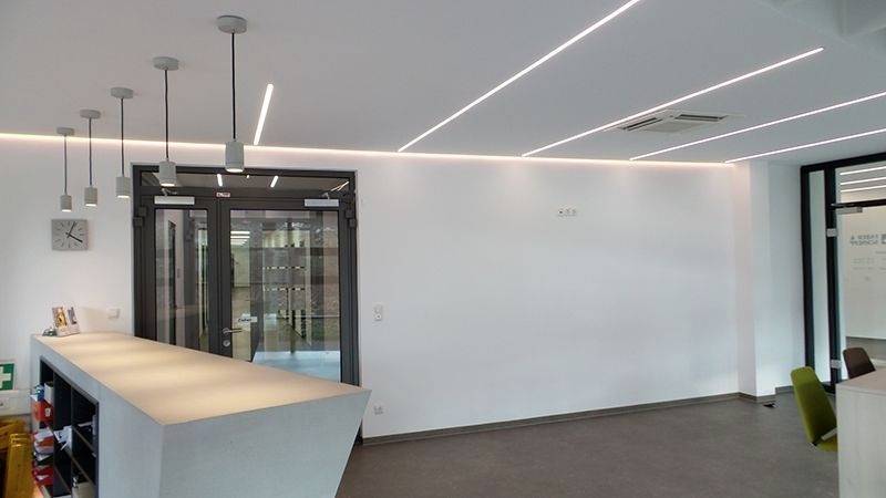 Moderner Büroraum mit hellem Empfangsbereich, minimalistischem Design und eingelassenen Deckenleuchten. Links Empfangstheke mit Hängelampen.
