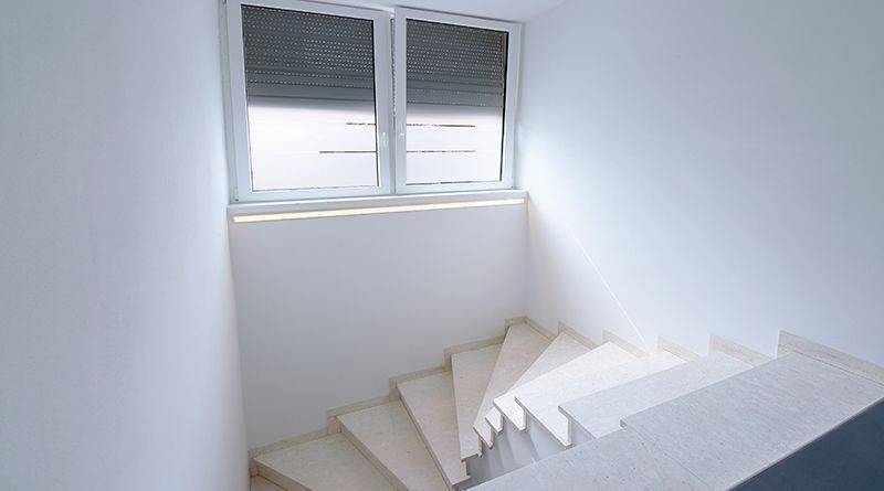 Eine weiße Treppe führt zu einem Fenster mit geschlossenen Rollos, das diffuses Licht in den Raum lässt. Minimalistisches Design.