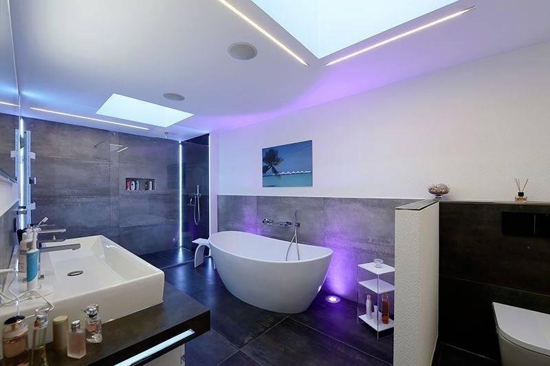 Modernes Badezimmer mit freistehender Badewanne, zwei Waschbecken und LED-Beleuchtung. Dunkle Fliesen und Deckenfenster sorgen für eine elegante Atmosphäre.