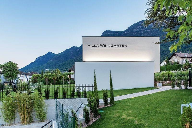 Eine moderne, weiße Villa mit der Aufschrift 'VILLA WEINGARTEN' steht in einer grünen Landschaft vor einem Gebirgshintergrund.