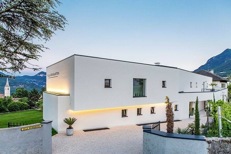 Modernes weißes Gebäude mit klaren Linien in einer Berglandschaft. Im Hintergrund eine Kirche und zypressenartige Pflanzen im Hof.