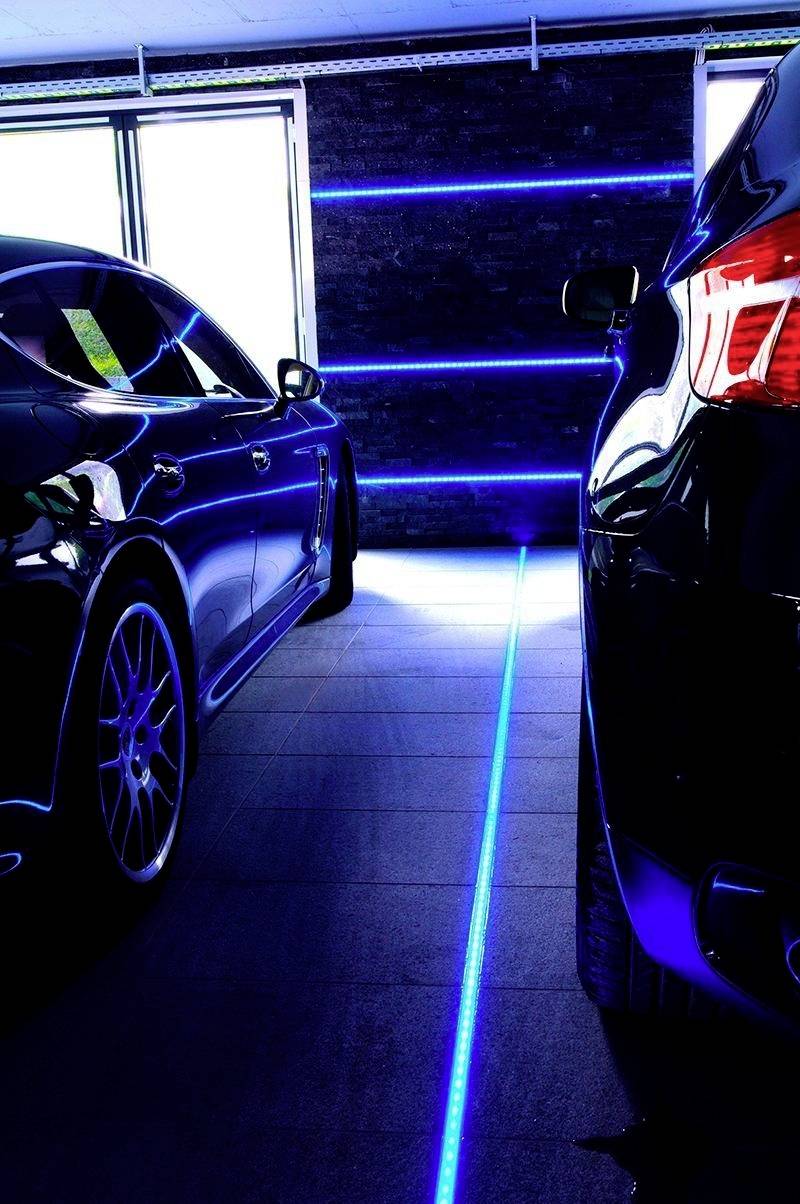 Zwei Autos in einer Garage mit blauer LED-Leiste am Boden zwischen ihnen, die den Parkplatz klar markiert. Hintergrund: schwarze Wand mit blauen Lichtern.