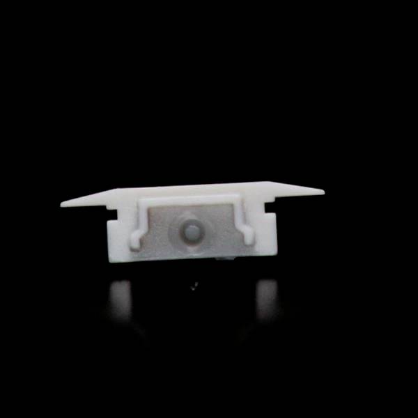 Ein kleines, weißes Kunststoffteil mit zwei seitlichen Armen, zentralem rundem Element, vor schwarzem Hintergrund.