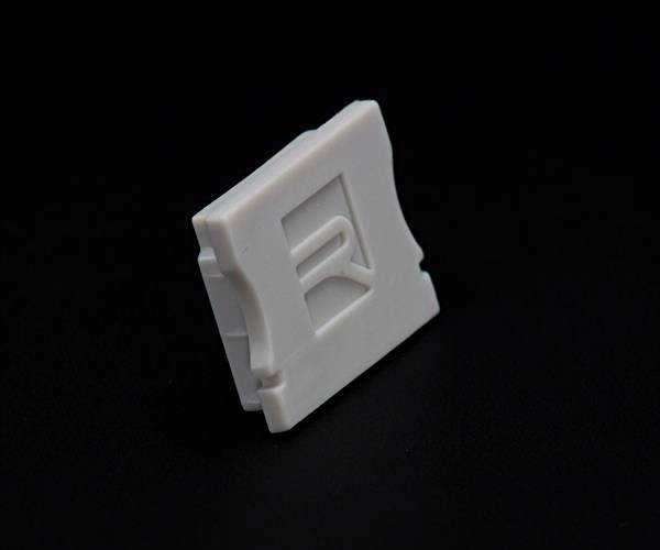Ein kleiner, weißer Kunststoffclip mit einem Reliefbuchstaben 'R' auf schwarzem Hintergrund.