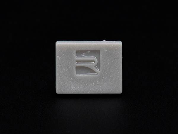 Eine rechteckige, weiße Taste mit einem vertieften, stilisierten 'R'-Logo auf schwarzem Hintergrund.