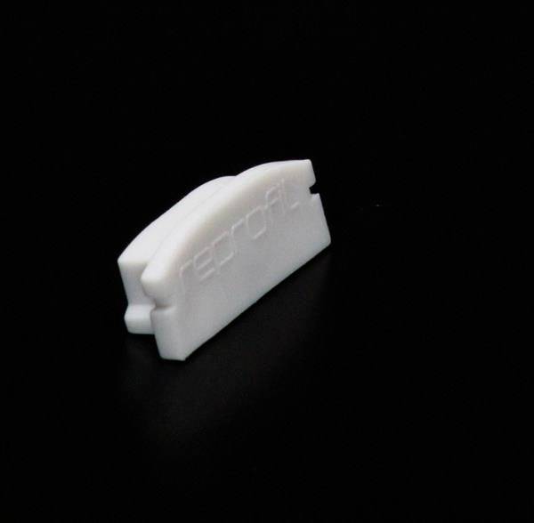 Ein weißes Kunststoffobjekt auf schwarzem Hintergrund, das wie eine kleine, abgerundete Lasche geformt ist.