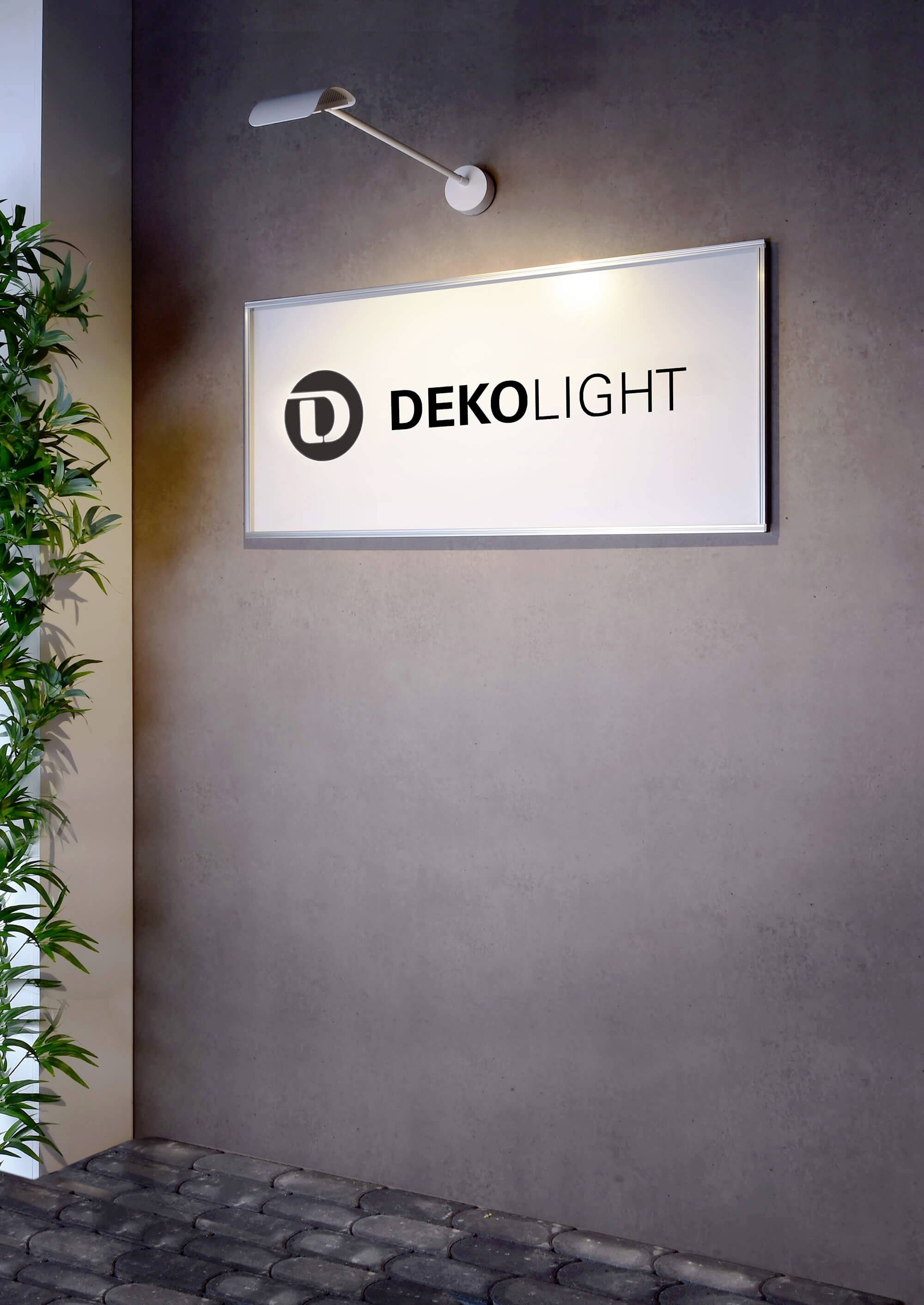 Ein Firmenschild mit der Aufschrift 'DEKOLIGHT' hängt an einer beleuchteten grauen Wand. Links ist ein Teil einer Pflanze sichtbar.