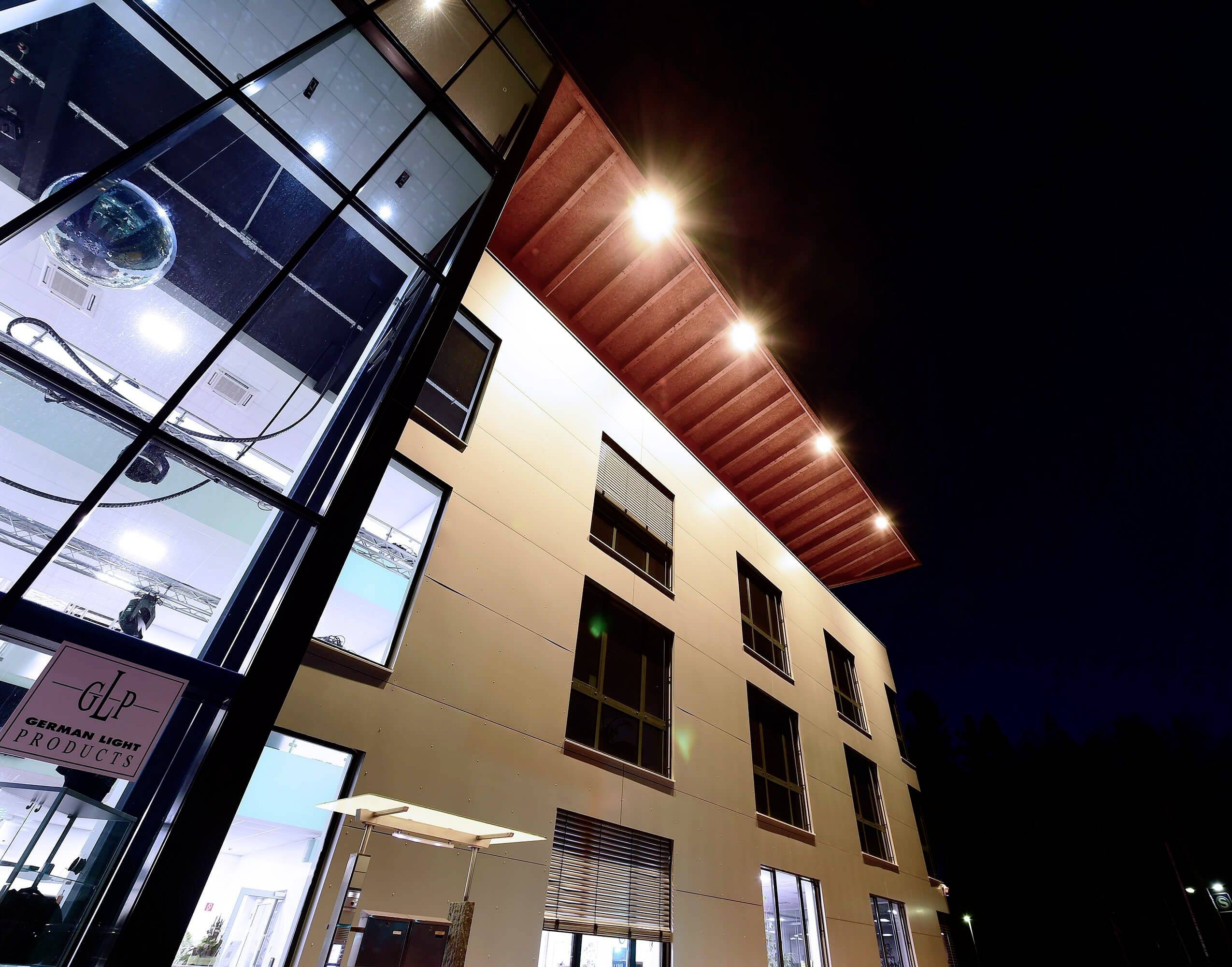 Das Bild zeigt ein modernes Gebäude bei Nacht. Es hat eine Glasfassade mit erleuchteten Fenstern und eine markante, rot beleuchtete Dachkante.