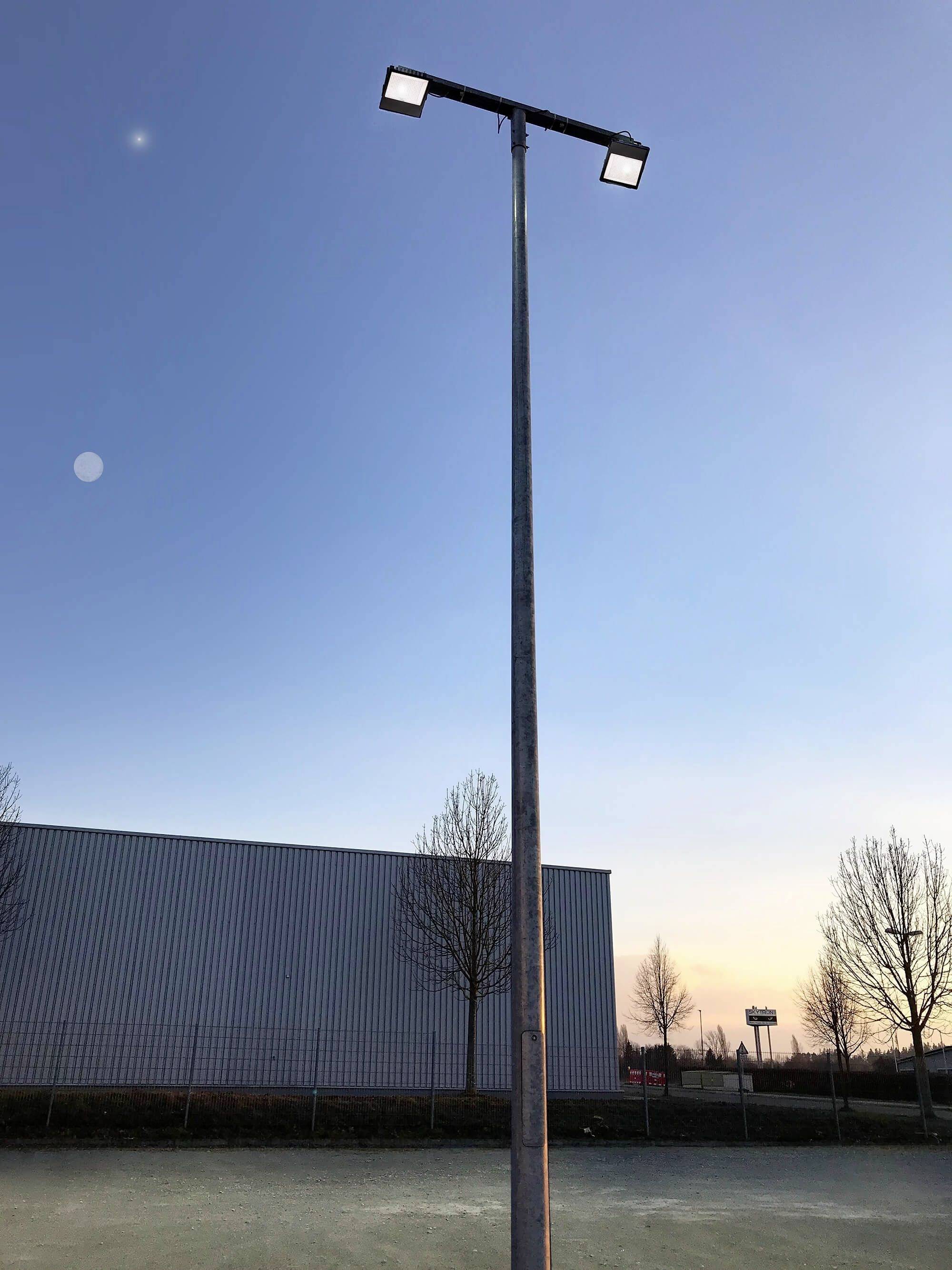 Eine hohe Straßenlaterne steht auf einem Platz neben einem modernen Gebäude. Der Himmel ist bei Dämmerung klar mit wenigen Wolken.