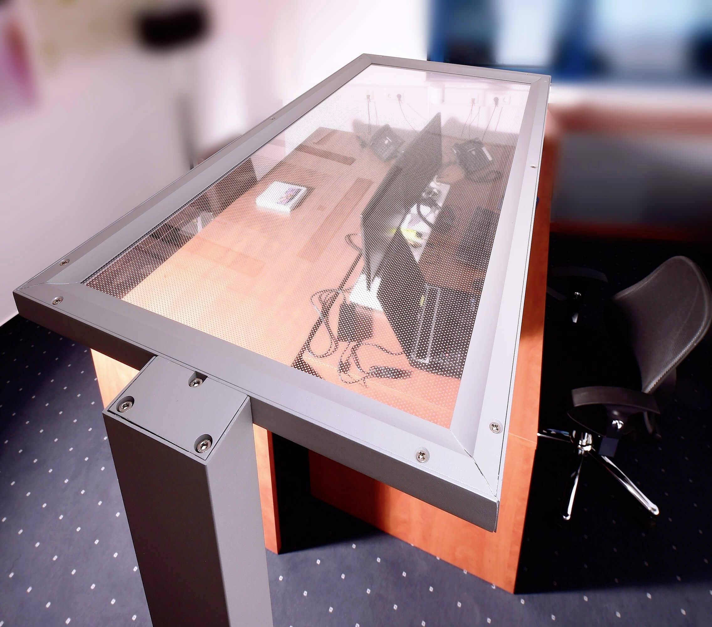 Ein schwebendes, modernes Pult mit Glasplatte zeigt darunter liegende Computerbildschirme und Kabel. Ein schwarzer Bürostuhl steht daneben.