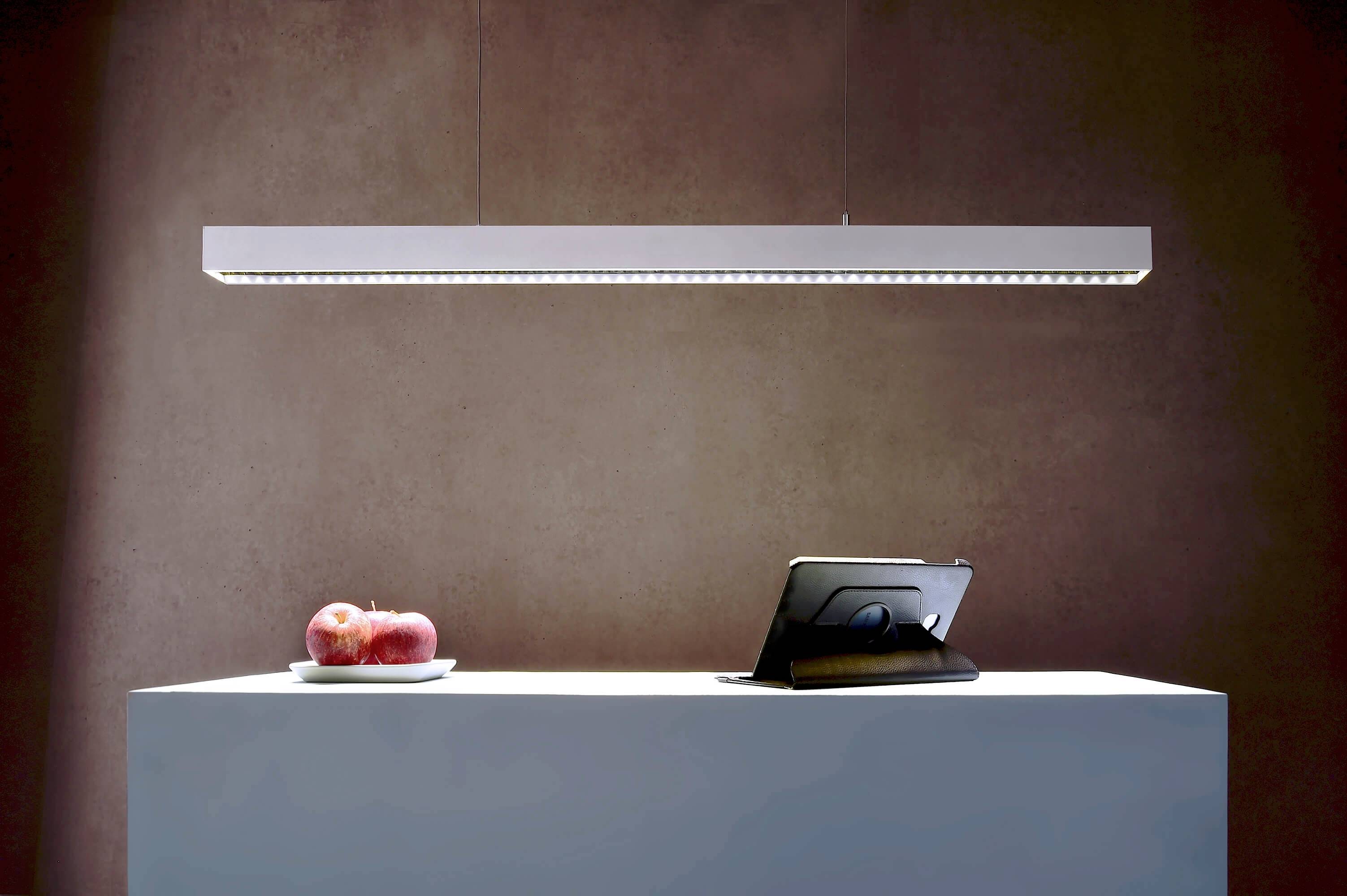 Ein moderner, rechteckiger Hängeleuchter leuchtet über einem Tisch mit einem Teller roter Äpfel und einem aufgestellten Tablet.