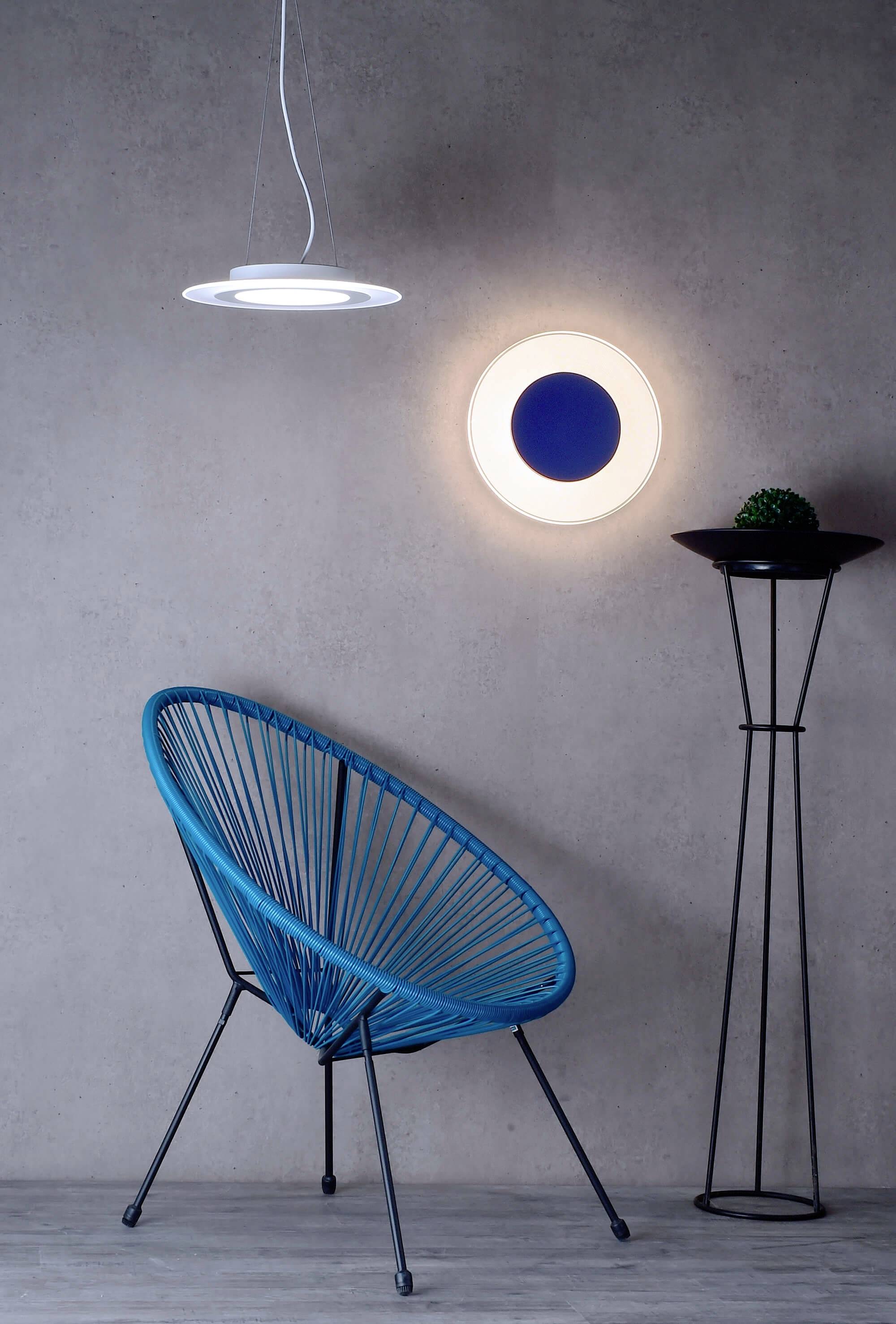 Ein blauer Stuhl mit dreieckigem Design steht vor einer grauen Wand. Rechts ist eine moderne Lampe an der Wand befestigt.