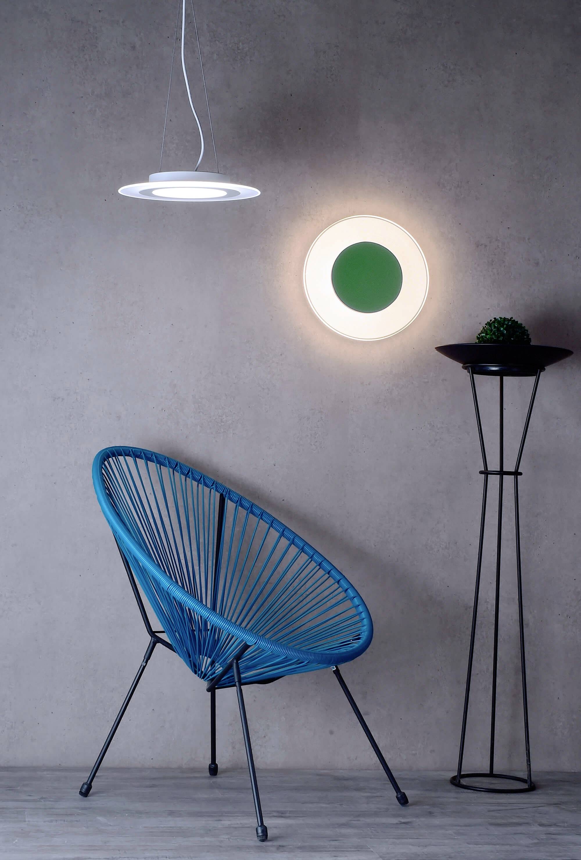 Ein blauer, runder Stuhl steht vor einer grauen Wand. An der Wand hängt eine runde Lampe, daneben ein hoher schwarzer Ständer mit Dekokugel.