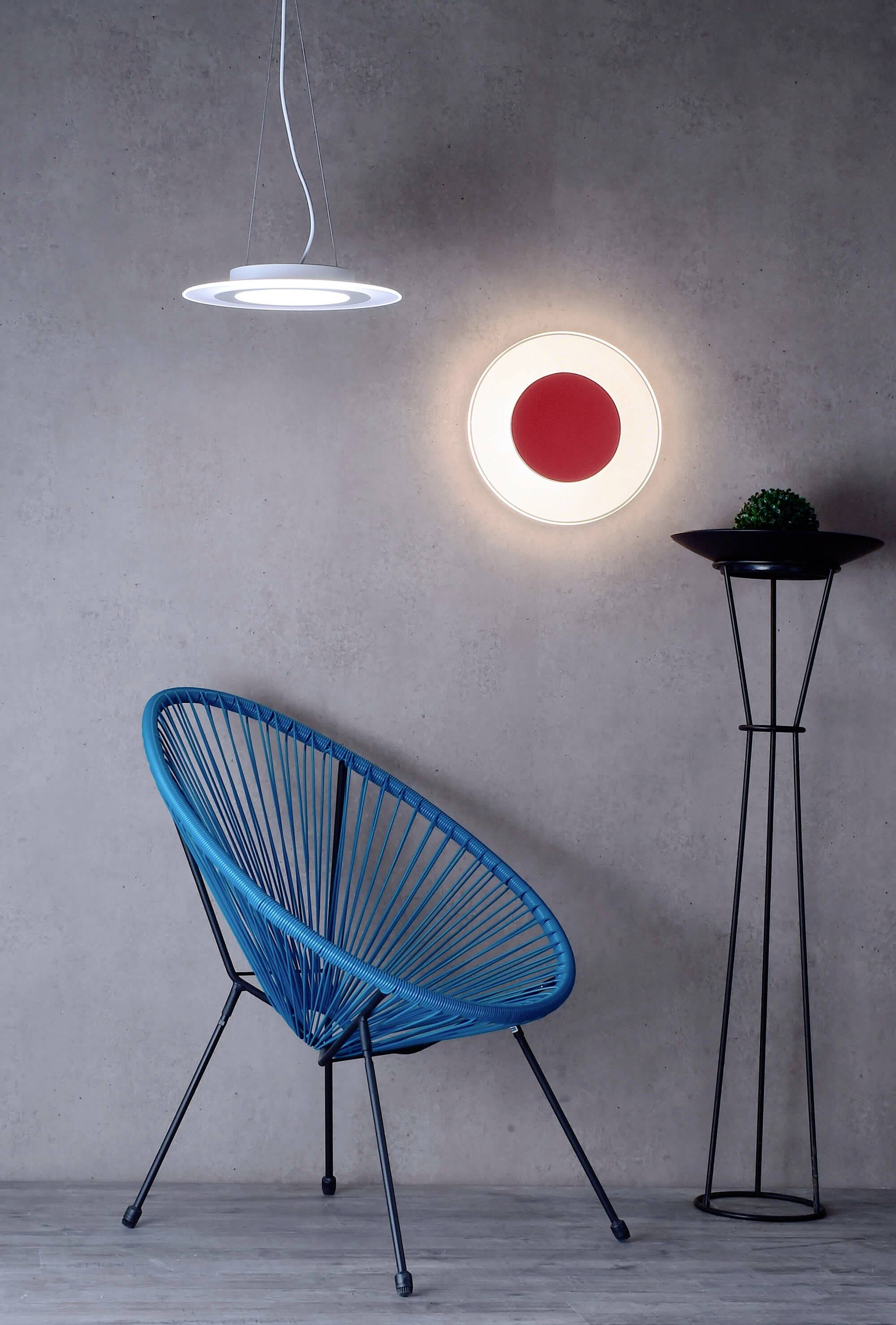 Ein blauer Stuhl vor einer grauen Wand, auf der eine runde Lampe mit rotem Zentrum leuchtet. Daneben steht ein hoher Pflanzenständer.