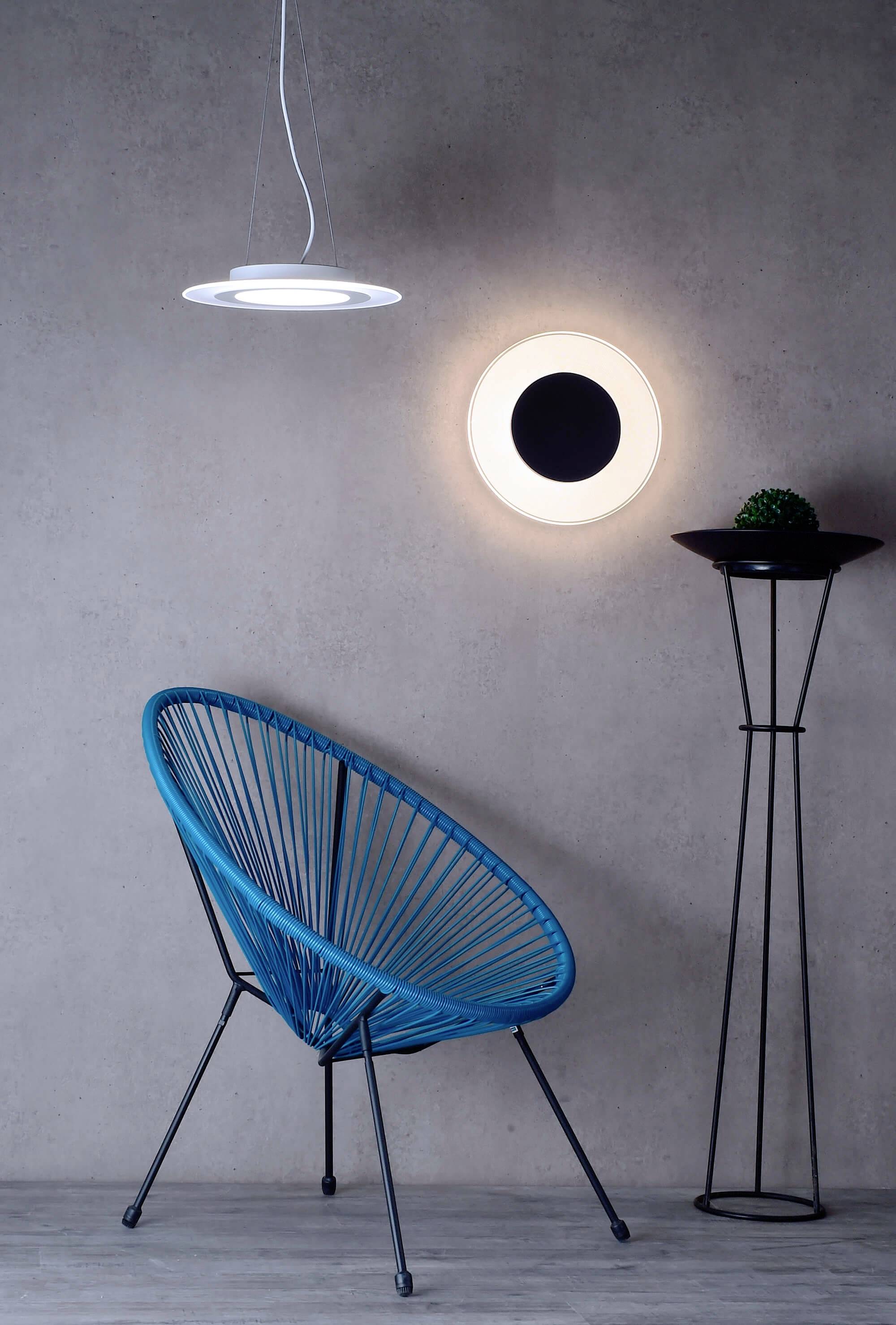 Ein blauer Stuhl steht neben einer modernen Lampe vor einer grauen Betonwand. Rechts steht ein hoher, schwarzer Ständer mit Pflanze.