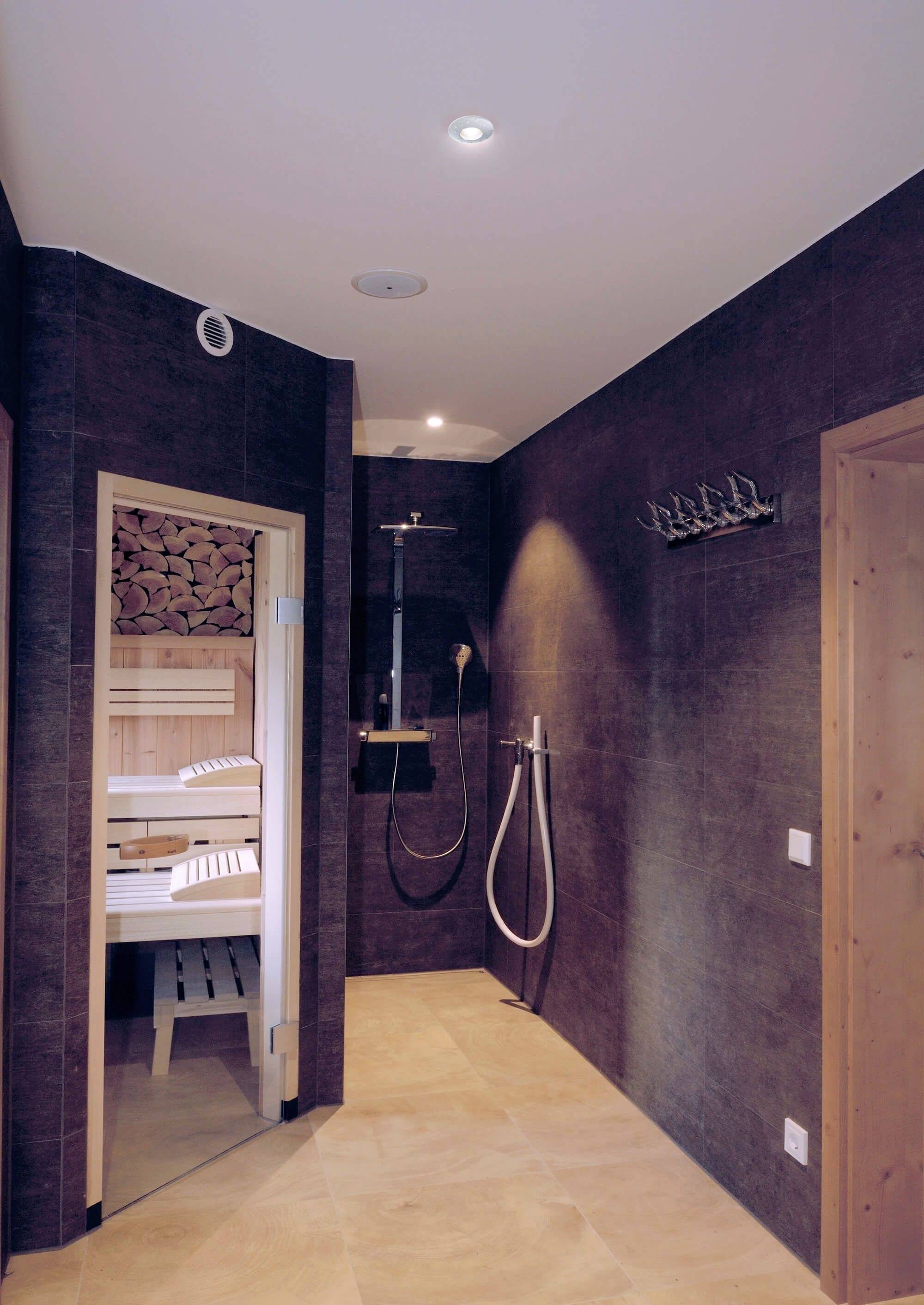 Dusche und Sauna in modernem Badebereich mit dunklen Wänden und hellem Boden. Links ist ein Stapel Brennholz zu sehen.