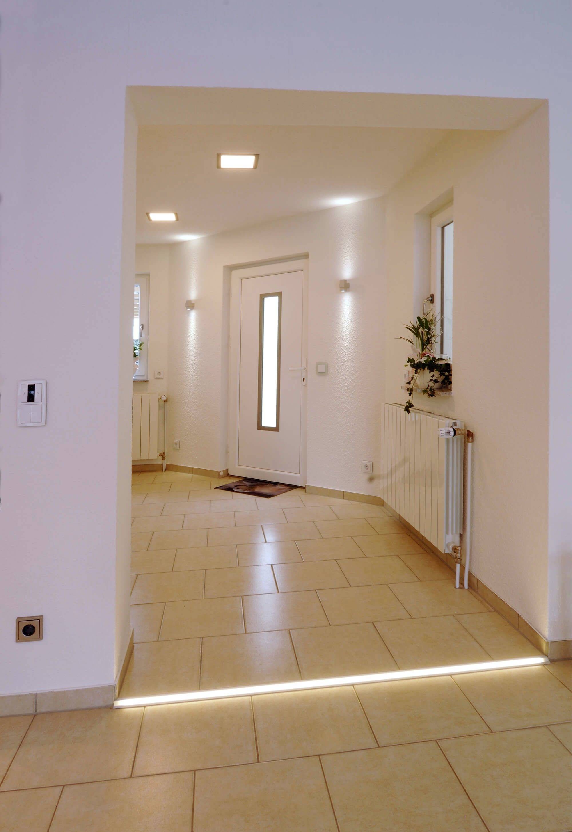 Eingangsbereich mit heller Fliese, weißer Tür und modernem Beleuchtungskonzept. Pflanzen am Fensterbrett neben Heizkörper.