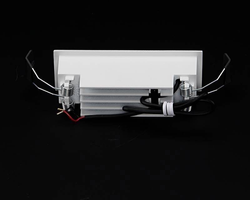 Rechteckige weiße Deckenleuchte mit abstehenden Metallclips an den Seiten und einer schwarzen Kabelverbindung auf schwarzem Hintergrund.