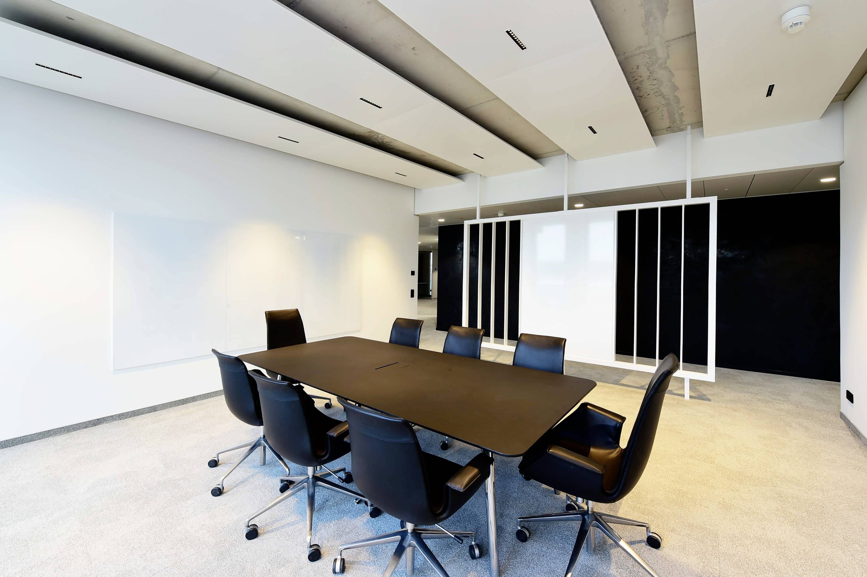 Ein moderner, leerer Konferenzraum mit einem großen schwarzen Tisch und acht schwarzen Stühlen auf hellem Teppichboden.