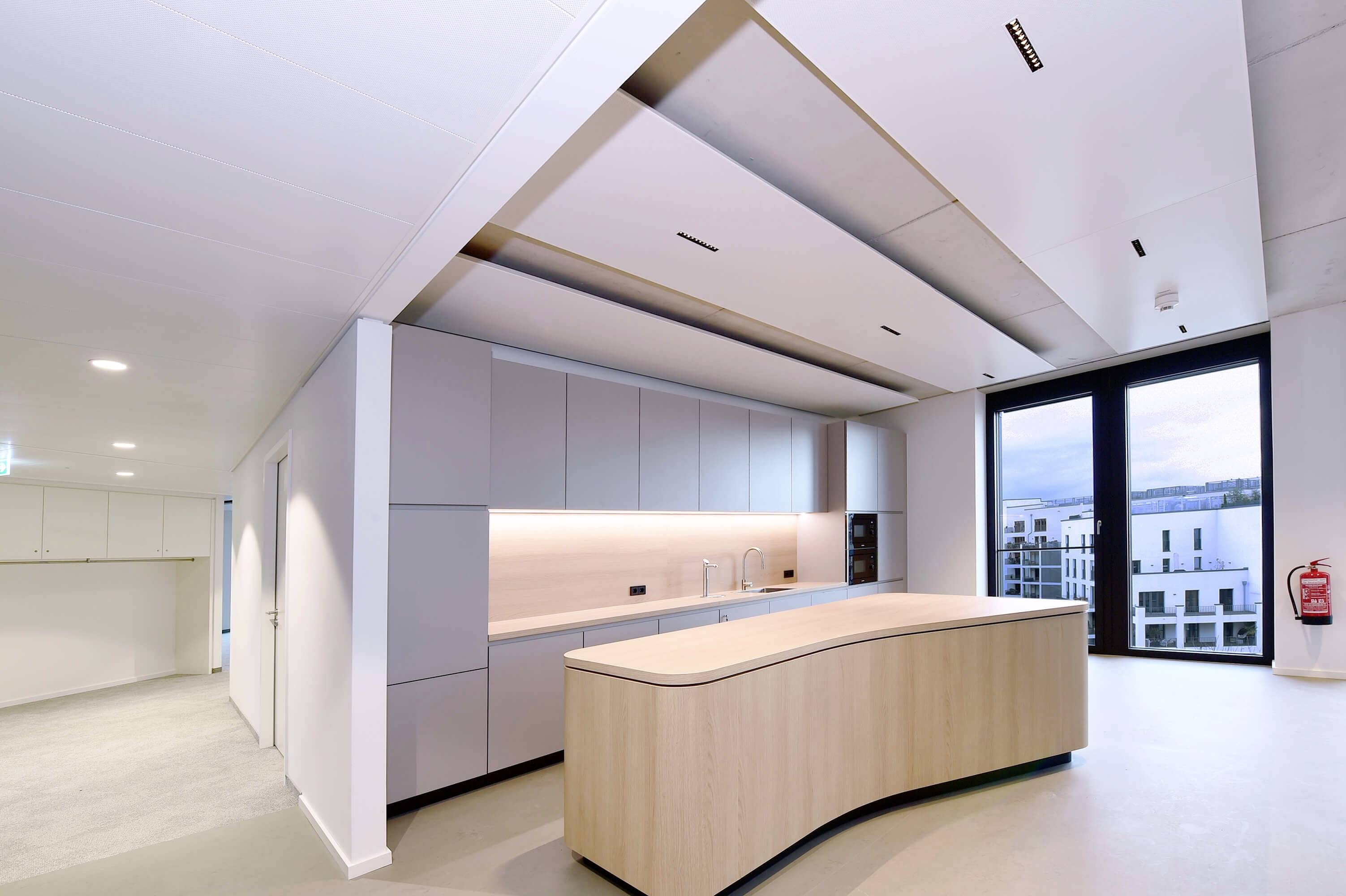 Moderne Küche mit einer geschwungenen Holzinsel, hellen Einbauschränken und großer Fensterfront mit Blick auf ein Gebäude im Hintergrund.