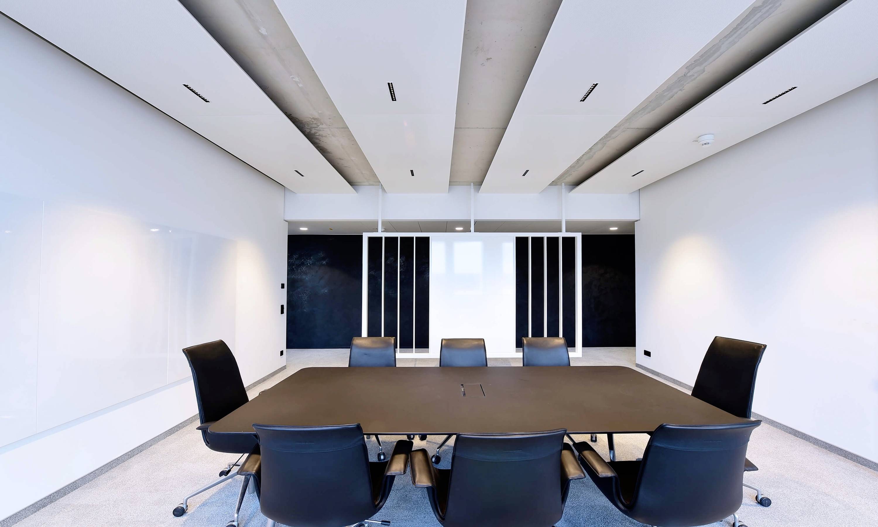 Konferenzraum mit großem Tisch und sieben Stühlen. Die Wände sind weiß, mit schwarzen Tafeln an der Stirnseite. Raum wirkt modern und minimalistisch.
