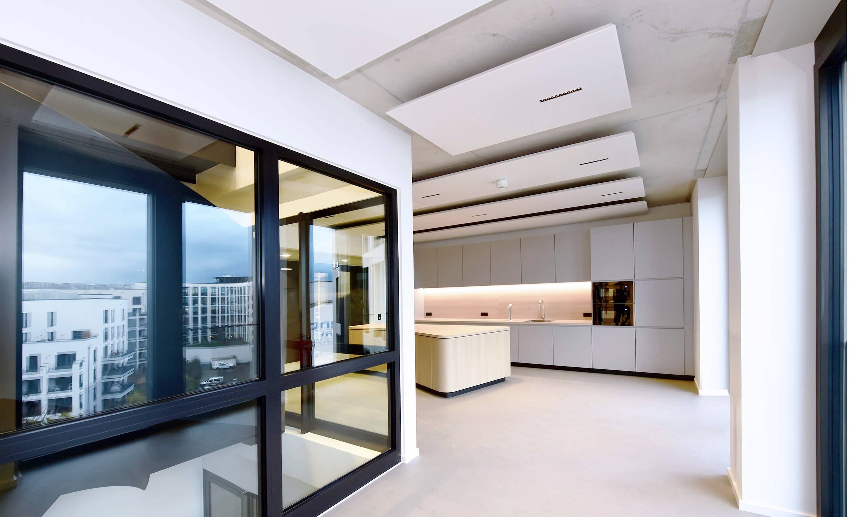 Moderne, helle Küche mit großen Fenstern, eingebautem Ofen und Mittelinsel in einem großzügigen Raum mit Blick auf Stadtgebäude.