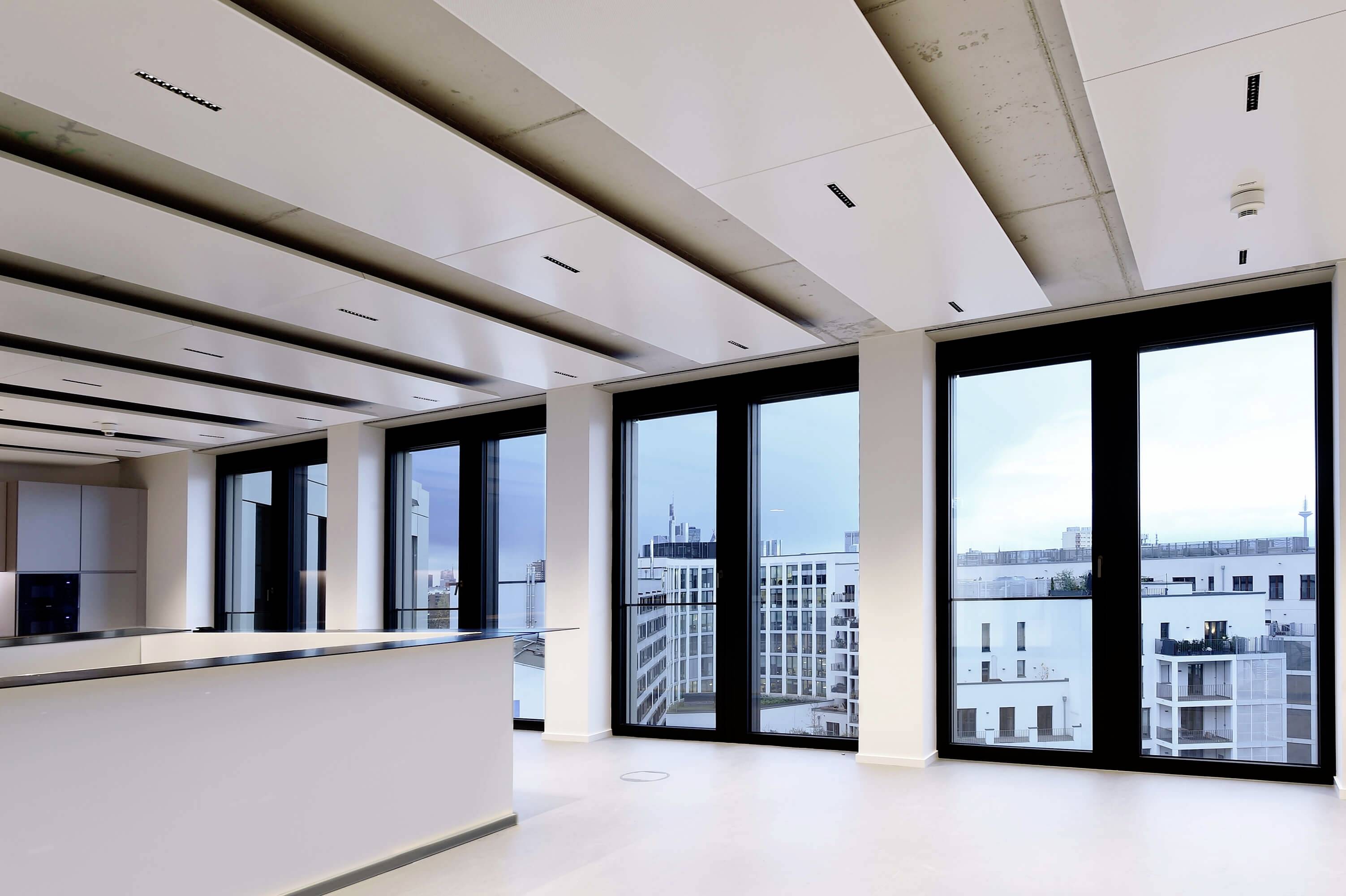 Ein modernes, leeres Büro mit großen Fenstern, die eine Aussicht auf Stadtgebäude bieten. Das Zimmer hat helle Wände und eine Decke mit integrierter Beleuchtung.