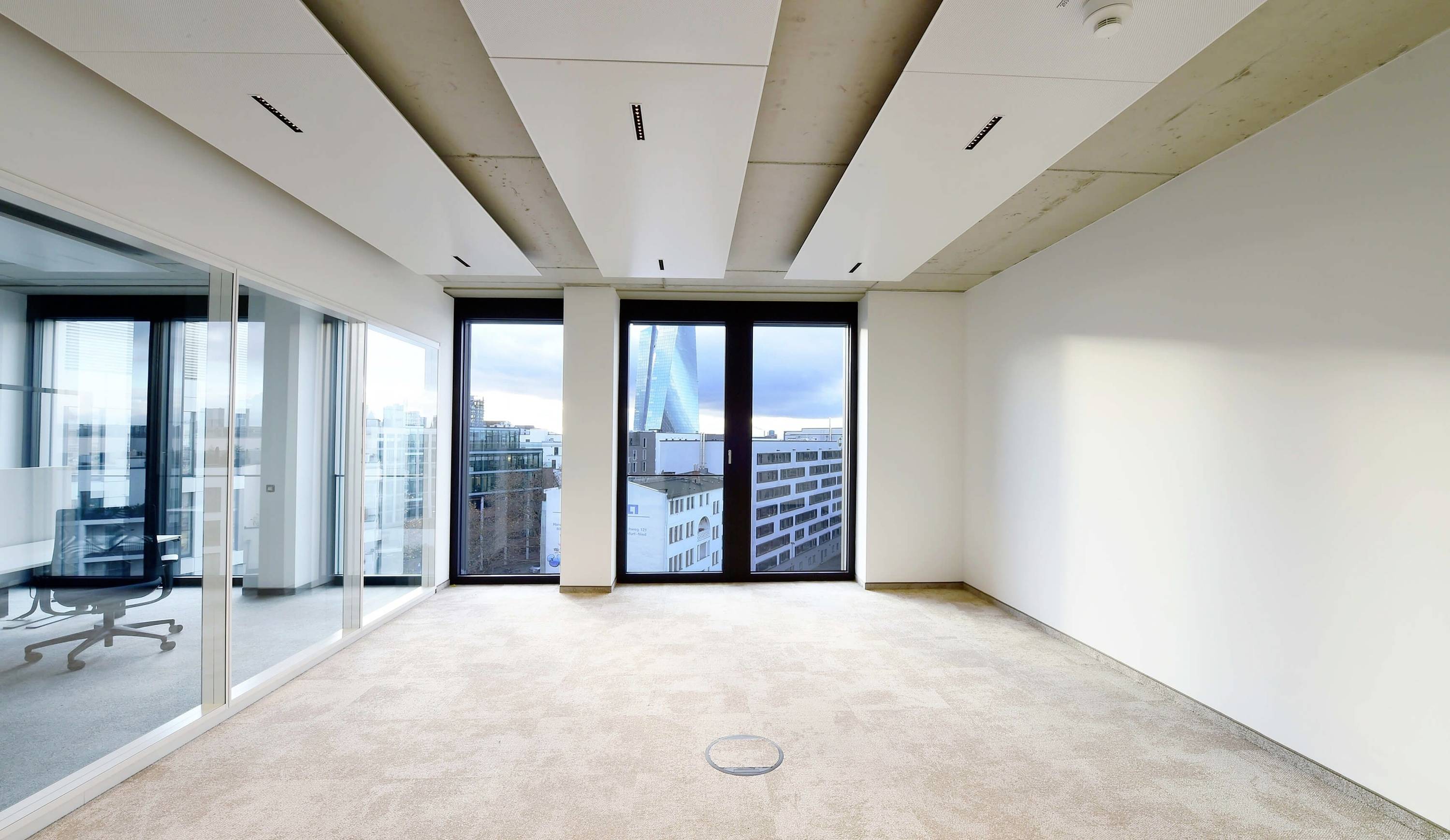 Leerer Büroraum mit Teppichboden, großen Fenstern und Stadtblick. Glaswände trennen einen Besprechungsbereich.