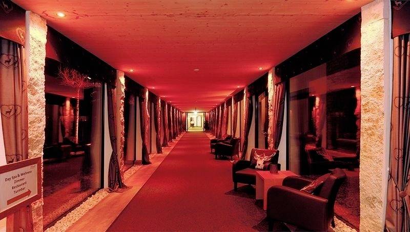 Ein langer, roter Flur mit Sofa und Sesseln an den Seiten, dekoriert mit Vorhängen und Steinen, führt zu einer hell erleuchteten Tür.