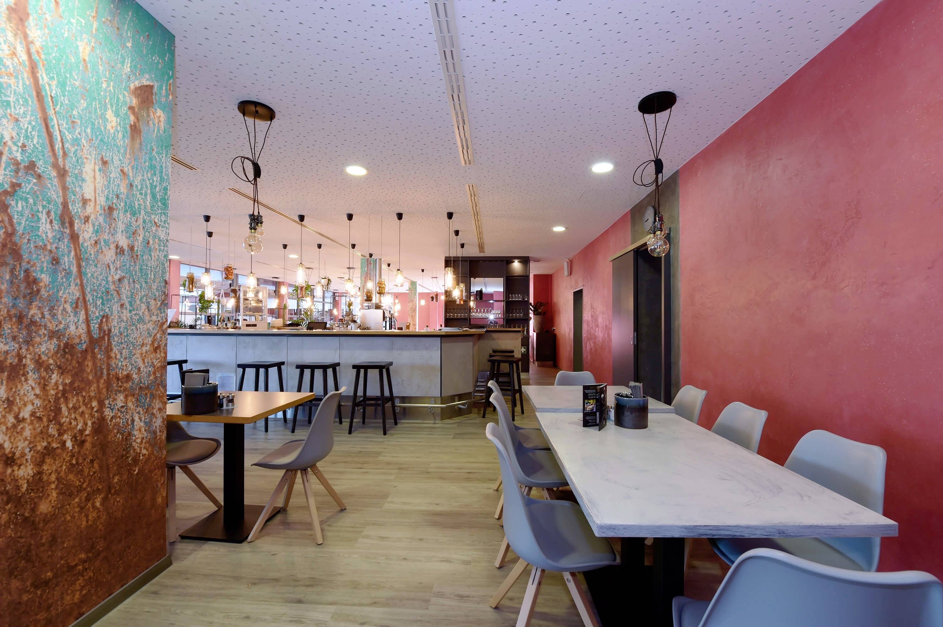 Ein modernes Café mit bunten Wänden, einer langen Bar, Sitzbereichen mit grauen Stühlen, stilvoller Beleuchtung und Holzboden.