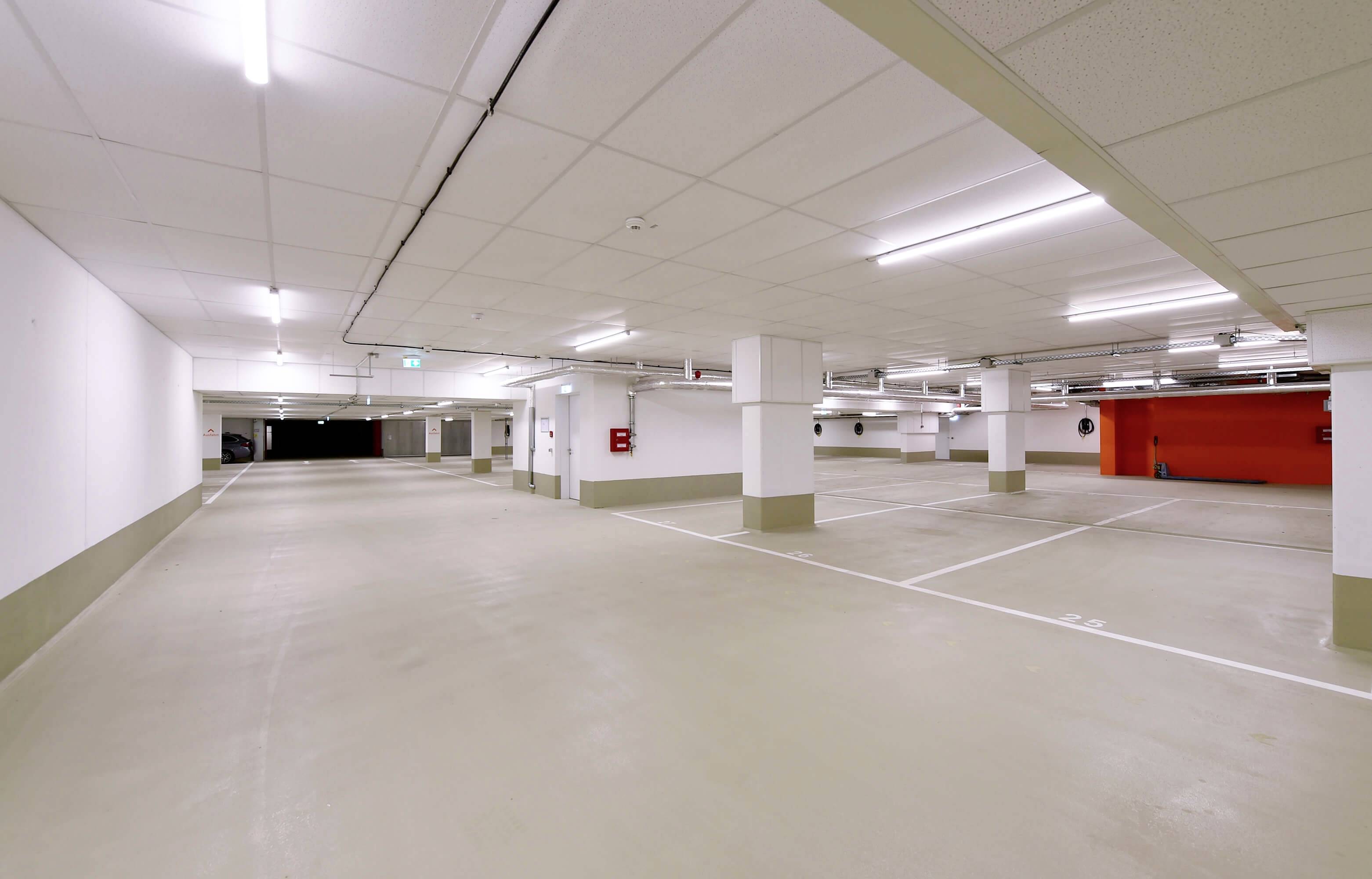 Ein leerer, gut beleuchteter Tiefgaragenparkplatz mit weißen Wänden und Markierungen auf dem Boden. An der Wand hängt ein Feuerlöscher.