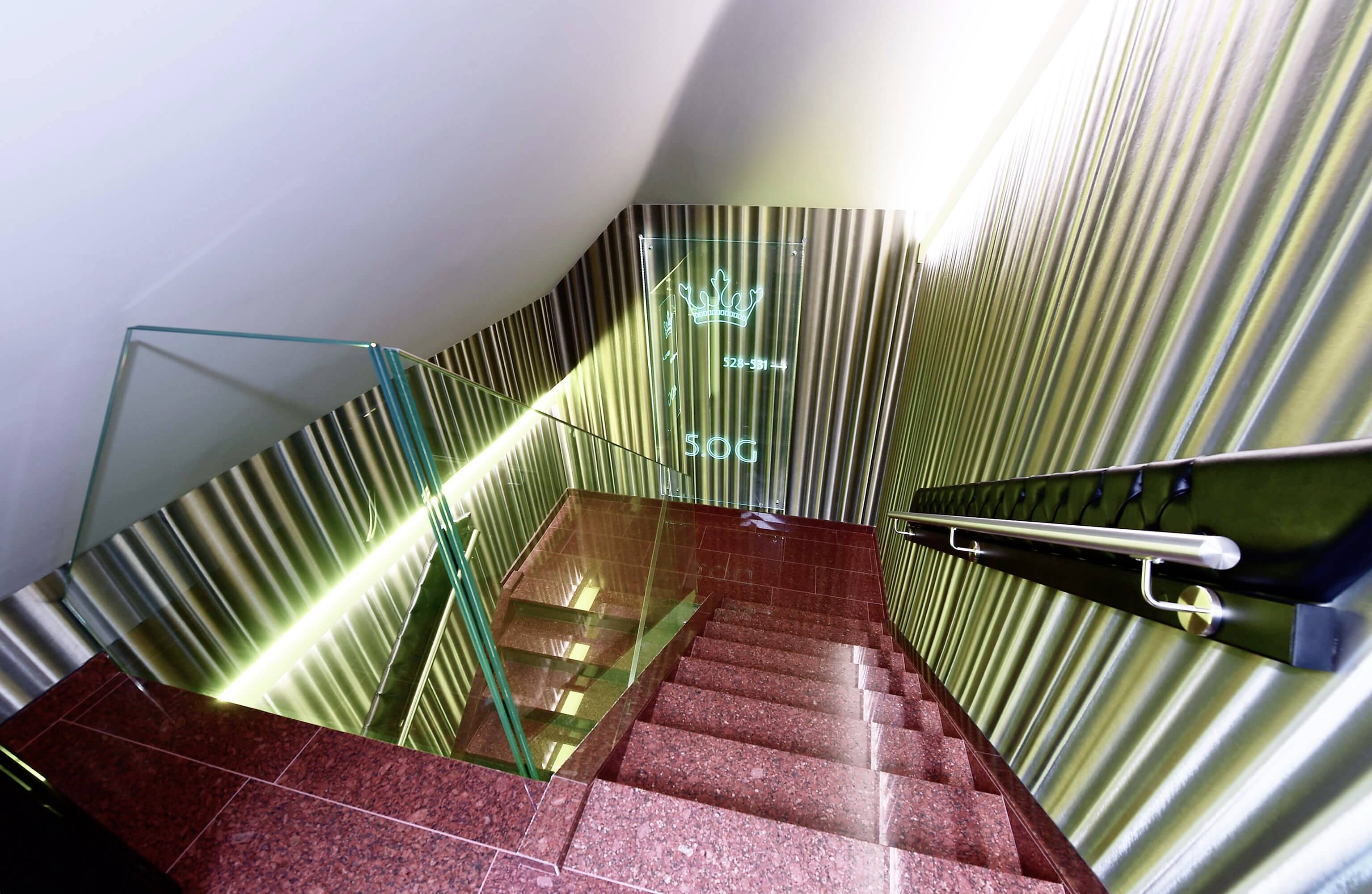 Rote Treppe in einem modernen Gebäude mit Glasgeländer und metallischen Wänden. Eine Krone und '500' sind an der Wand projiziert.