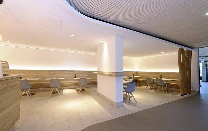 Heller, moderner Essbereich in einem Café mit Holztischen, grauen Stühlen und warmem Licht. Links dekorative Holzelemente.