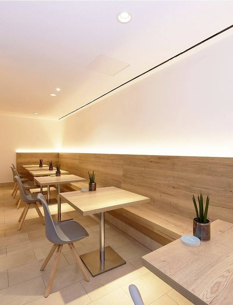 Moderne Cafeteria mit Holztischen und grauen Stühlen. Kleine Pflanzen als Dekoration. Warme Beleuchtung schafft eine gemütliche Atmosphäre.