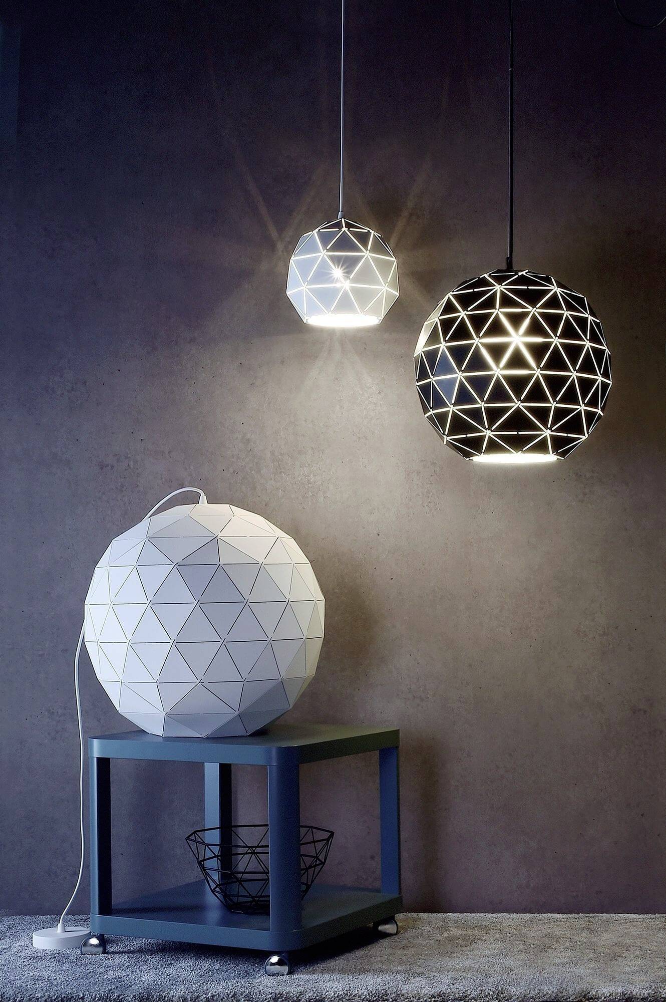 Drei moderne geometrische Deckenlampen in Weiß und Schwarz hängen vor einer grauen Wand. Eine Lampe steht auf einem Tisch darunter.