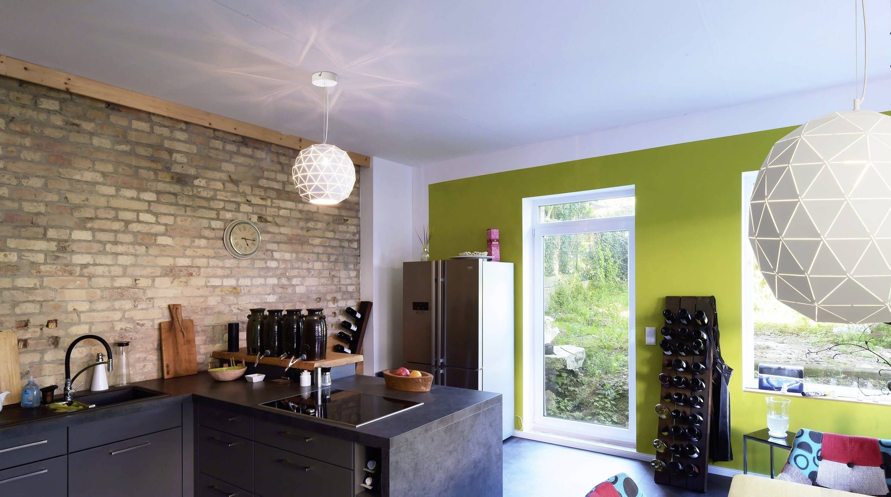Moderne Küche mit Backsteinwand, freistehendem Kühlschrank, Kochinsel und grüner Akzentwand. Große Fenster bieten Gartenblick.