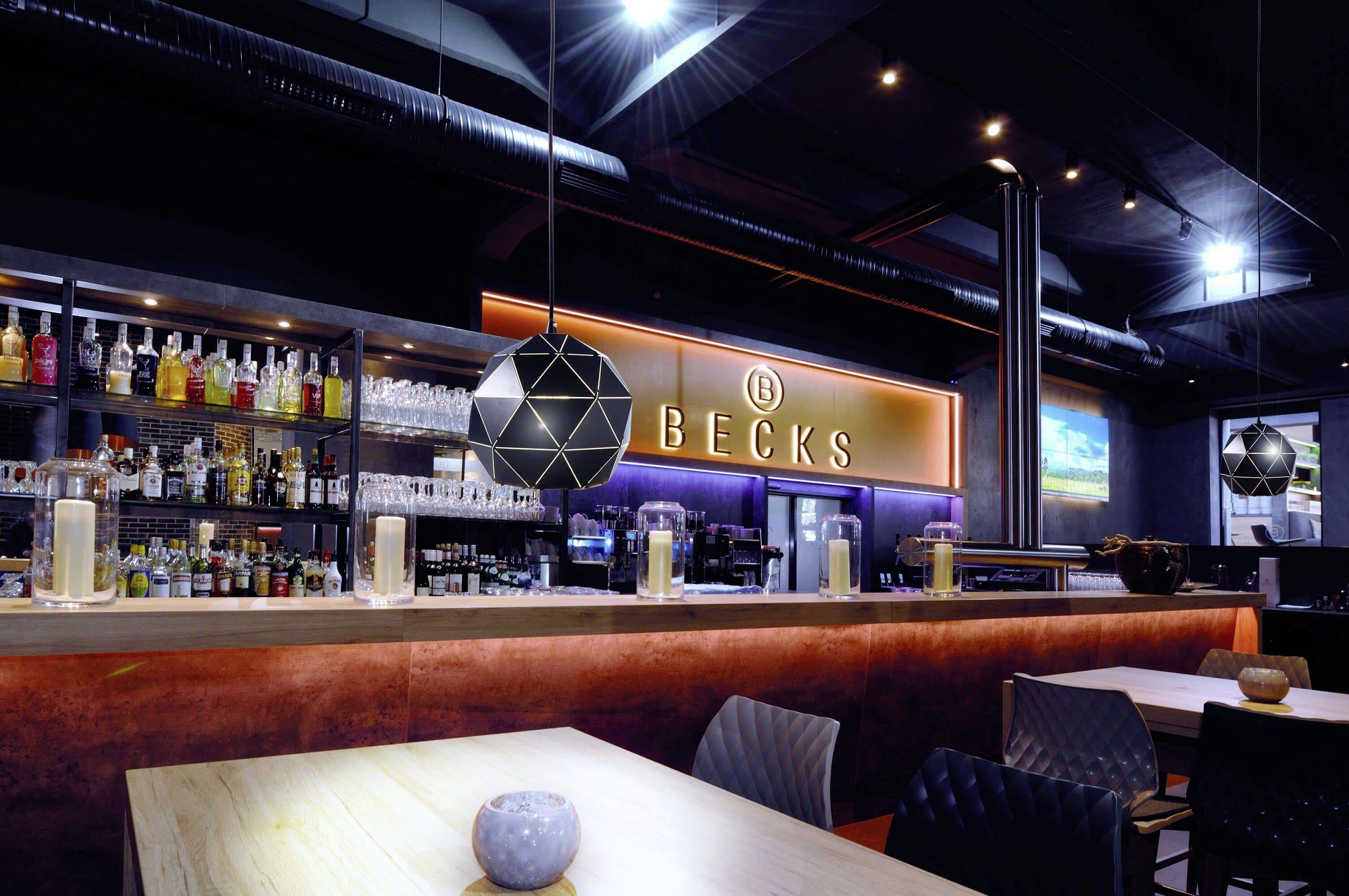 Moderne Bar mit beleuchteter Theke und Regalen voller Flaschen im Hintergrund. Neon-Schriftzug 'Becks' an der Wand.