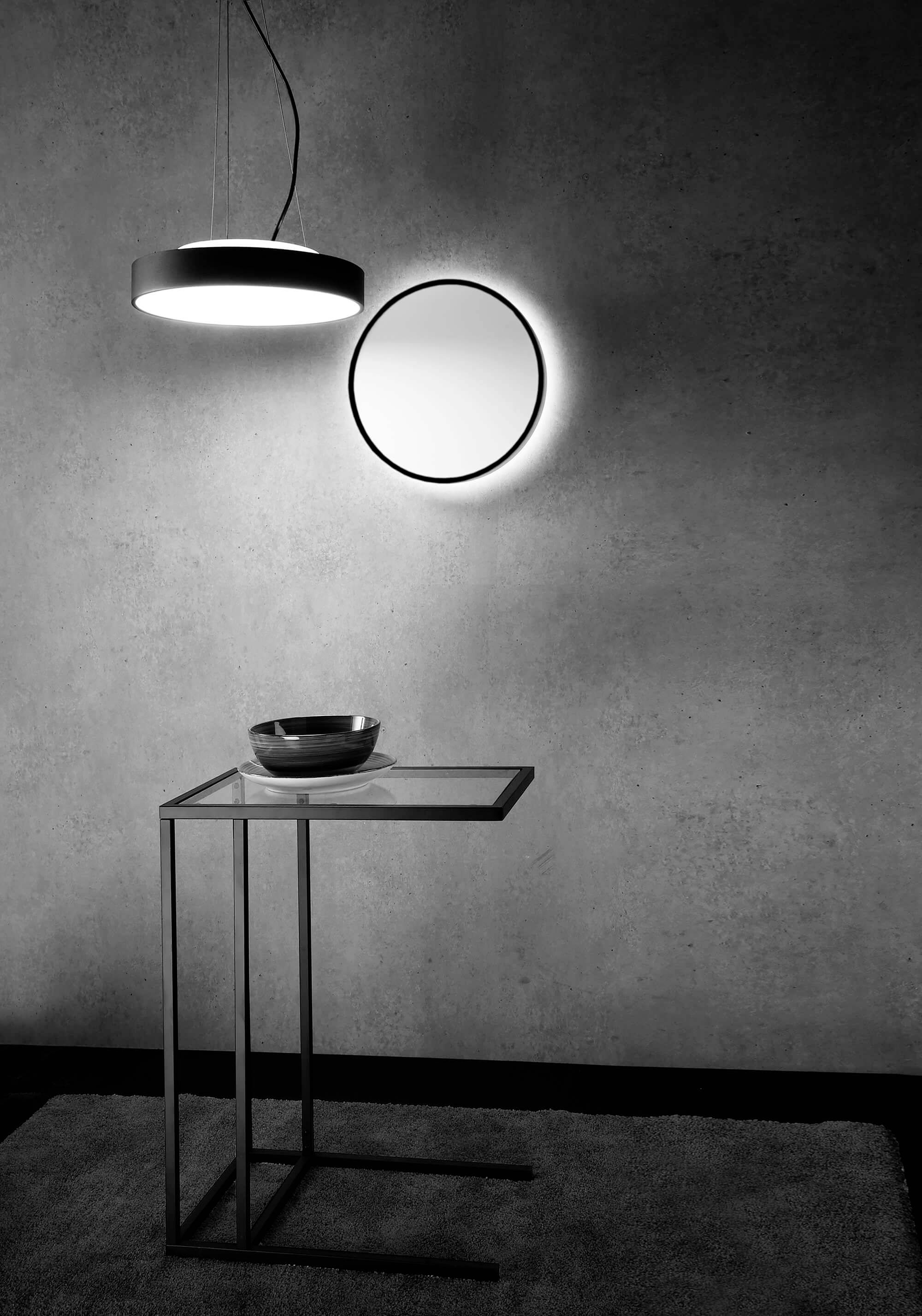 Ein minimalistischer Raum mit einem runden Spiegel, einer schwebenden Lampe und einem gläsernen Beistelltisch mit Schüssel darauf.
