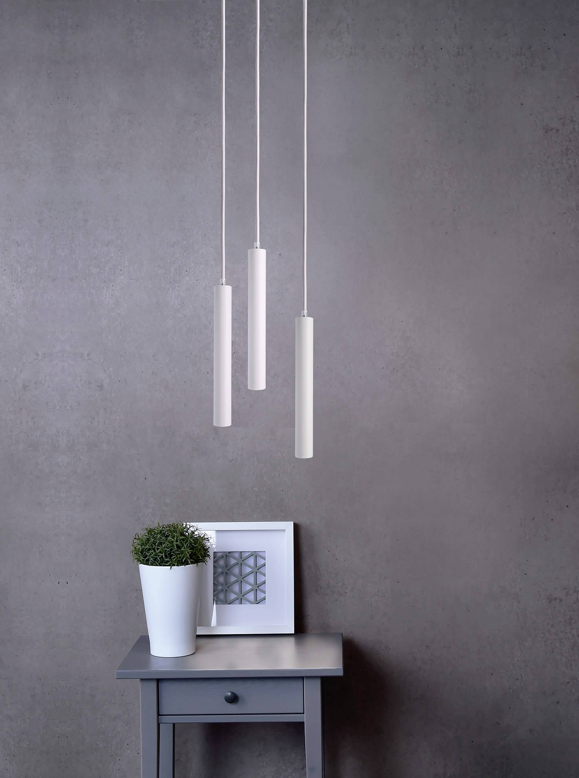 Minimalistische Zimmereinrichtung mit drei hängenden, zylindrischen Lampen über einem kleinen Tisch, der eine Pflanze und ein gerahmtes Bild trägt.