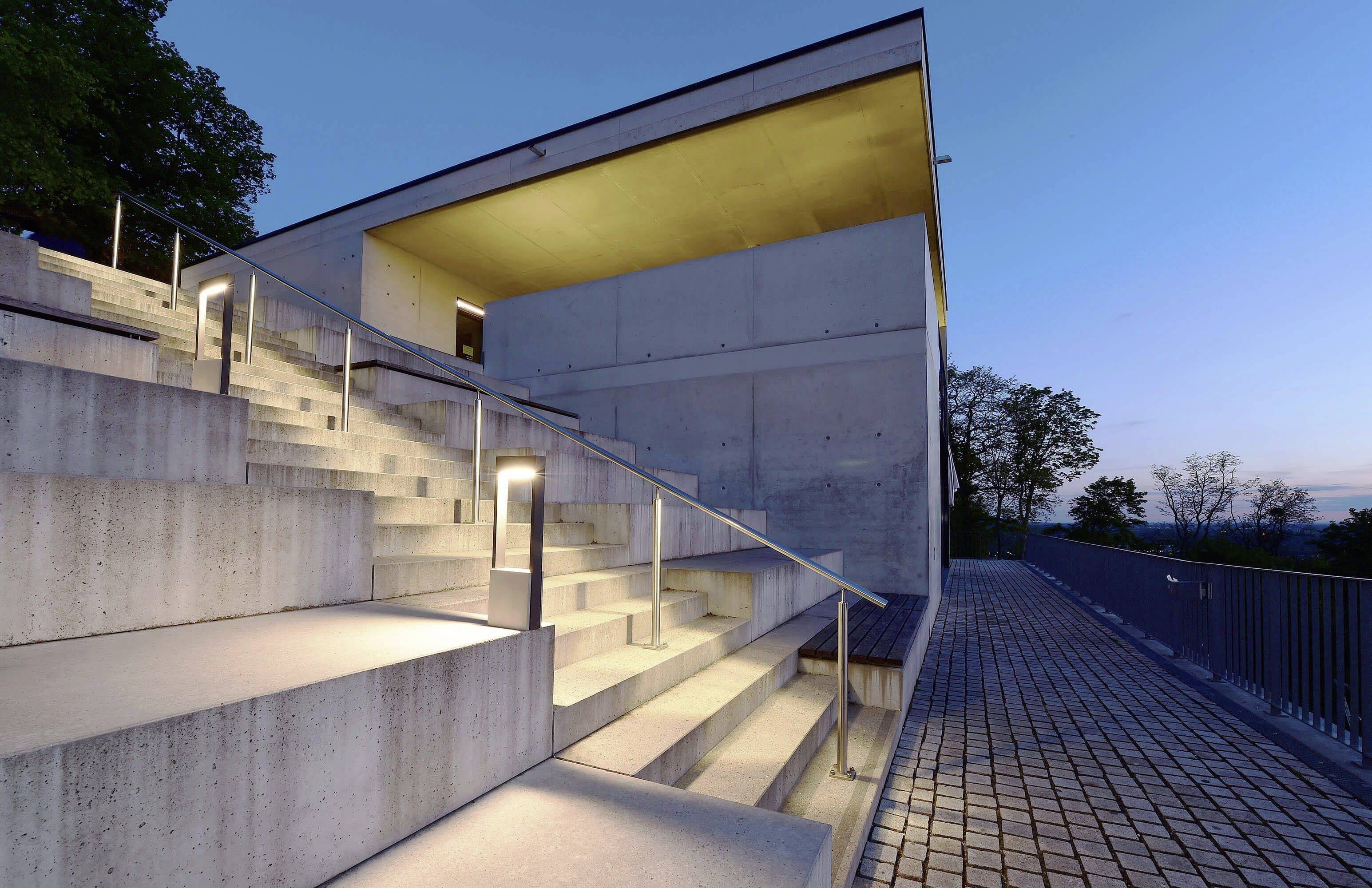 Ein modernes Gebäude mit beleuchteten Treppen und einem Geländer bei Dämmerung. Der geometrische Stil und die klare Architektur dominieren.