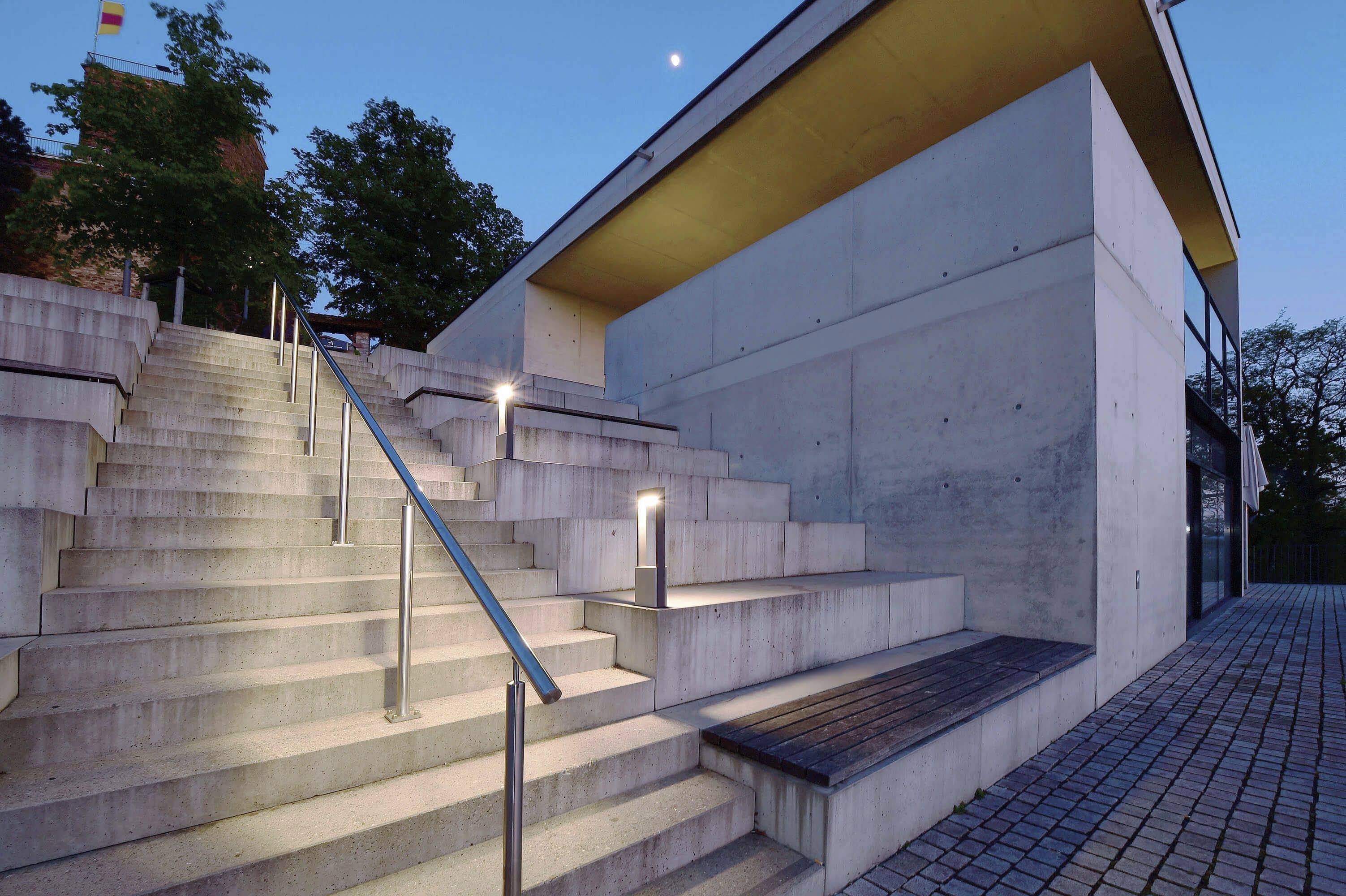 Ein moderner Gebäudezugang mit beleuchteten Betonstufen und Geländer, darunter ein gepflasterter Weg; umgeben von Bäumen bei Dämmerung.